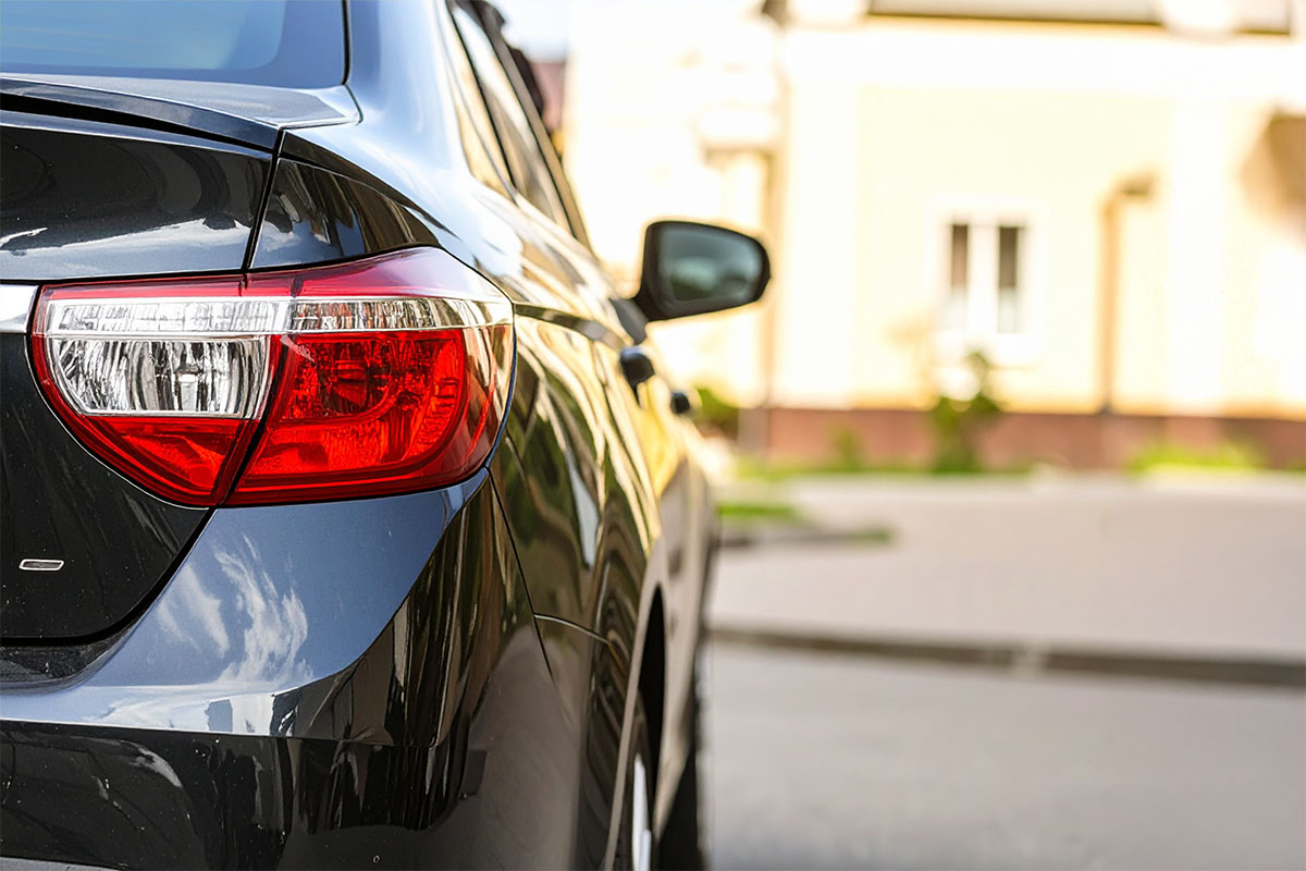 A close-up view of the rear of a dark-colored car, focusing on the red taillight, with a blurred background featuring a light-colored building and pavement.