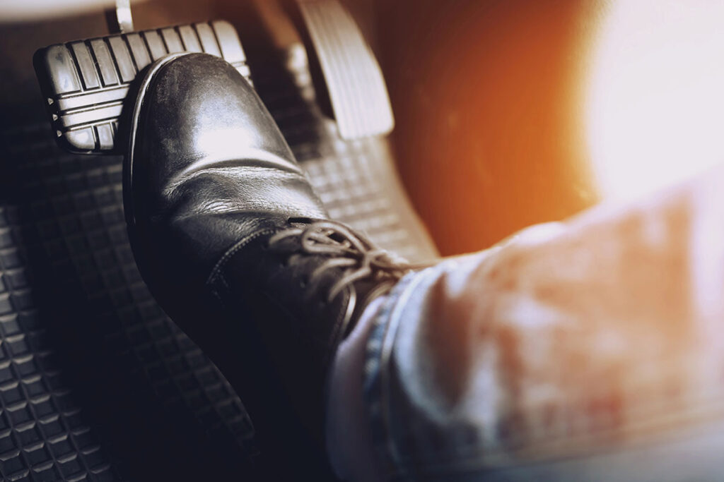 A close-up of a black shoe pressing down on a car brake pedal, with a blurred background showing part of a denim pant leg.