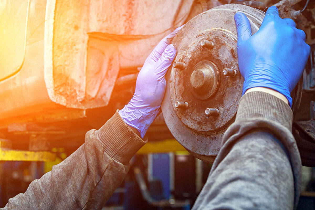 A close-up of two hands wearing blue gloves, working on a car brake disk in an automotive workshop. The brake disk appears worn and dusty, with tools and equipment visible in the background.