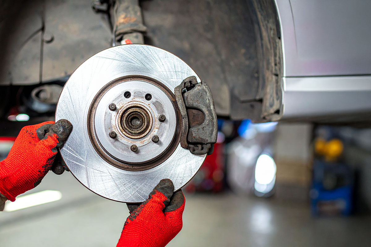 A close-up of hands wearing red gloves holding a silver brake disc, with a vehicle in the background, during a brake replacement or maintenance process.