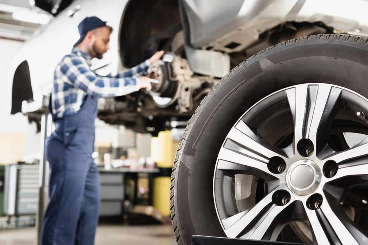 Reparing Wheel Hub A mechanic in blue overalls and a plaid shirt is inspecting a car's brake system, with a close-up view of a shiny alloy wheel in the foreground. The garage setting features tools and equipment in the background.