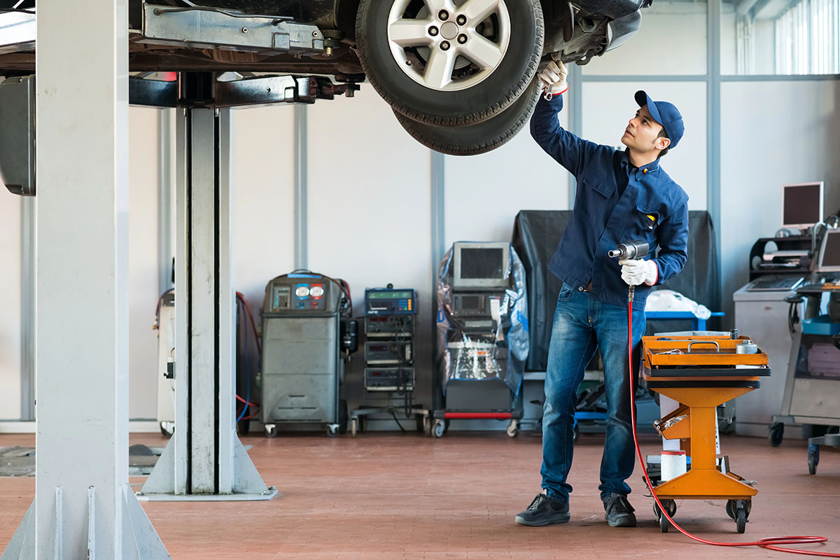 A mechanic in a blue uniform works under a raised vehicle in an auto repair shop, using a tool while standing next to an orange tool cart and various equipment in the background.
