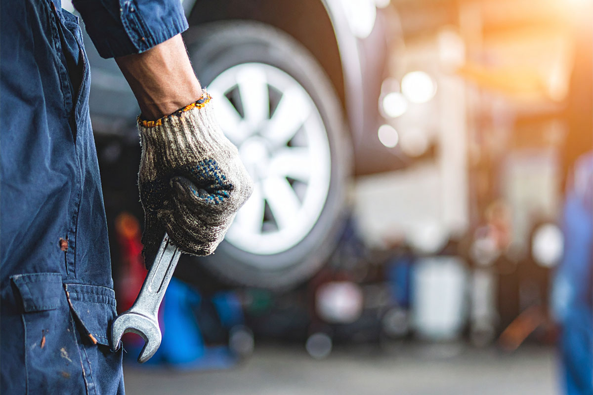 A mechanic holding a wrench in a garage, wearing a textured glove, with a blurred car wheel and tools in the background.