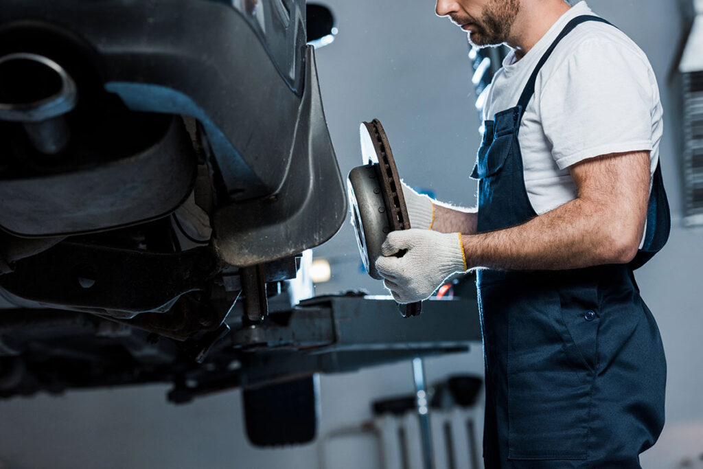 A mechanic in a blue jumpsuit and white gloves holds a brake disc while working under a lifted car in a garage.