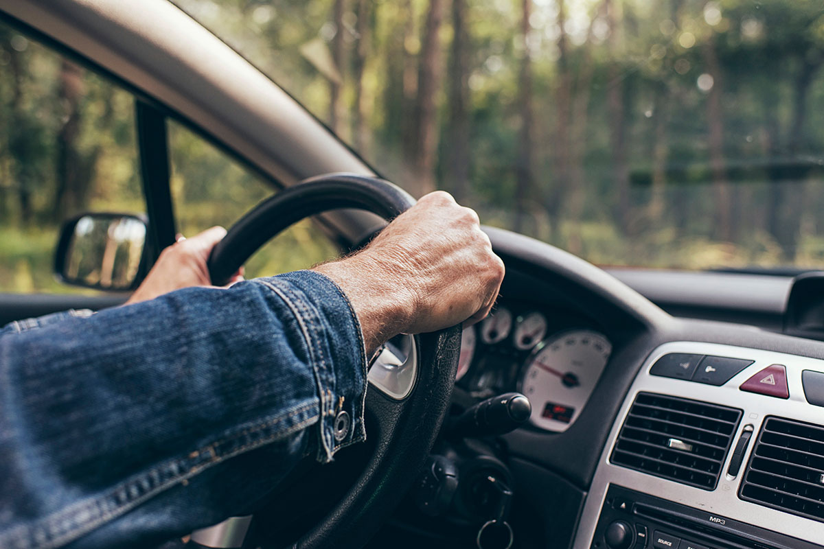 A close-up view of a person's hand gripping the steering wheel of a car, with a blurred background of trees and foliage visible through the windshield.