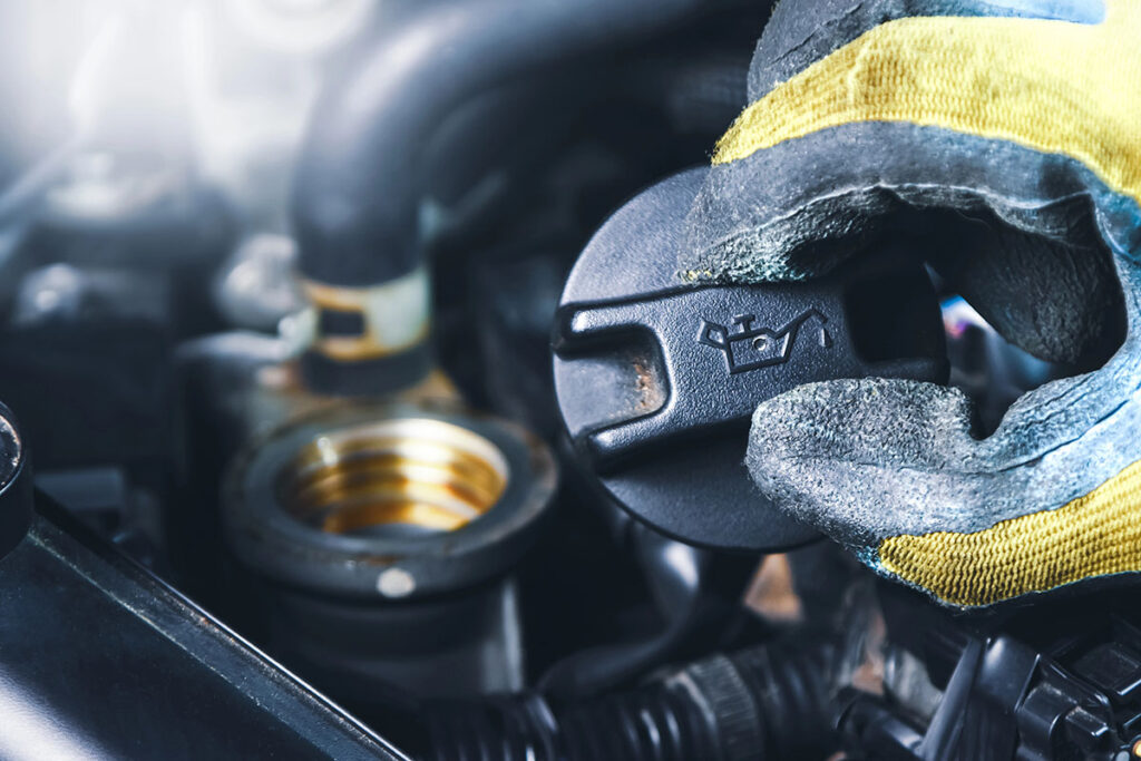 A gloved hand is holding an oil cap above an engine, with a focus on the cap that features an oil can icon, surrounded by various engine components.