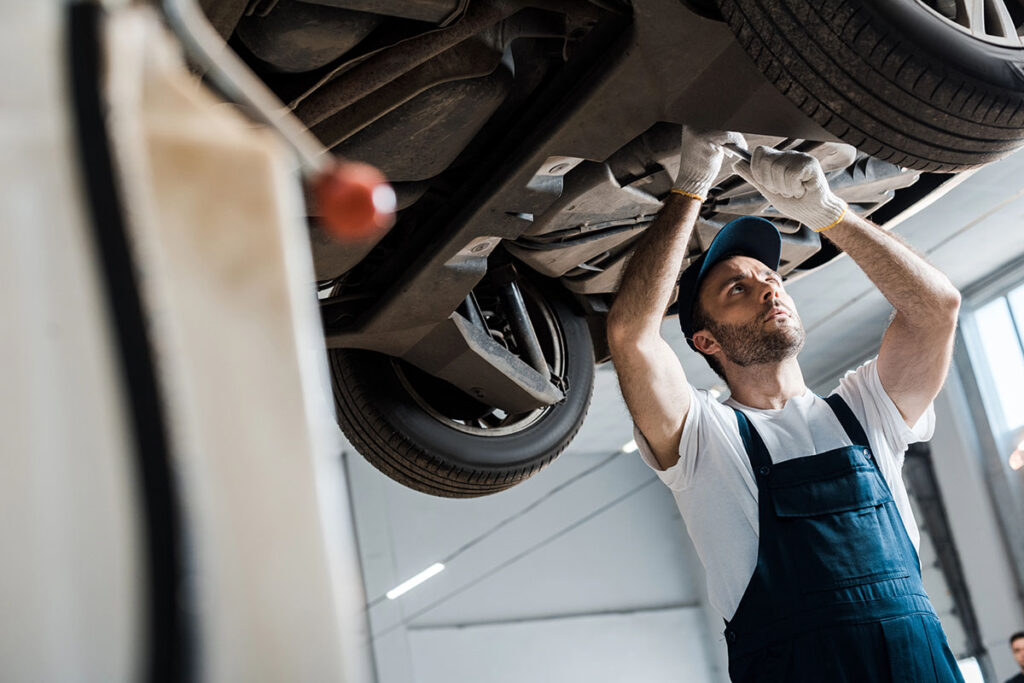 A mechanic wearing a blue cap and gloves is working underneath a car, focused on a repair while positioned on a service lift in a well-lit garage.