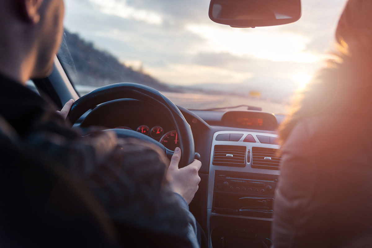 A driver holds the steering wheel of a car, with a passenger visible on the right side. The scene captures a sunset skyline through the windshield, illuminating the dashboard and interior.