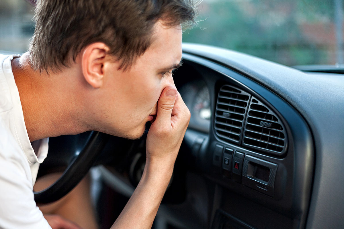 A man grimaces while sitting in a car, covering his nose with his hand, suggesting he is experiencing an unpleasant smell. The car's dashboard is visible in the foreground.