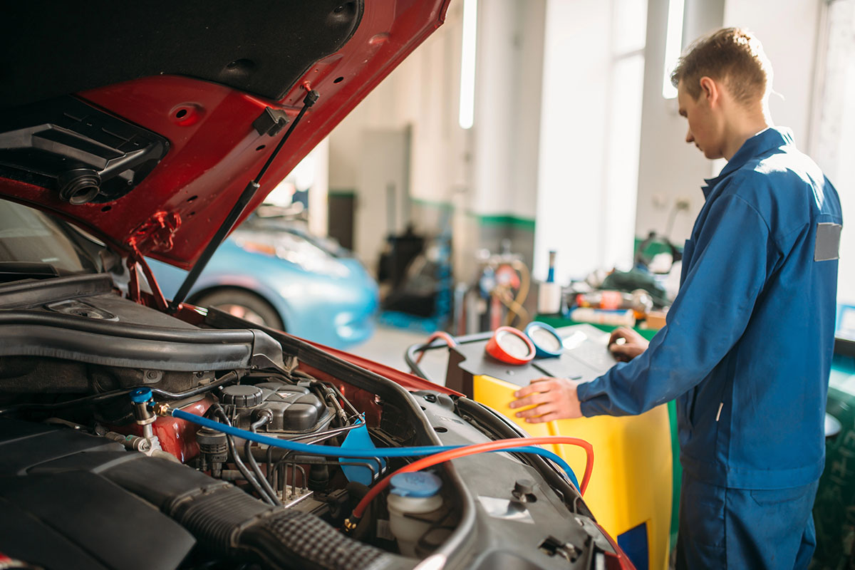 A mechanic in a blue jumpsuit works on a car engine with the hood raised, using diagnostic equipment located on a yellow machine nearby. A second car is visible in the background.