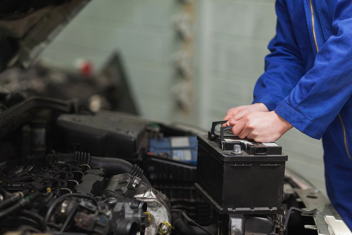 A mechanic in a blue jumpsuit is preparing to lift a car battery out of an engine compartment.