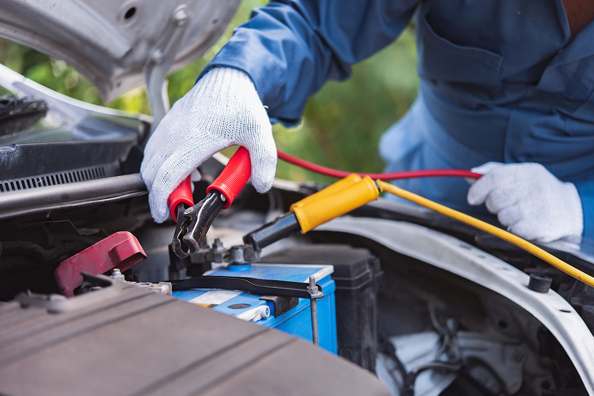 A person wearing a blue jumpsuit and white gloves is connecting jumper cables to a car battery under the hood of a vehicle. The jumper cables are red and yellow, with one clamp positioned on the positive terminal of the battery.