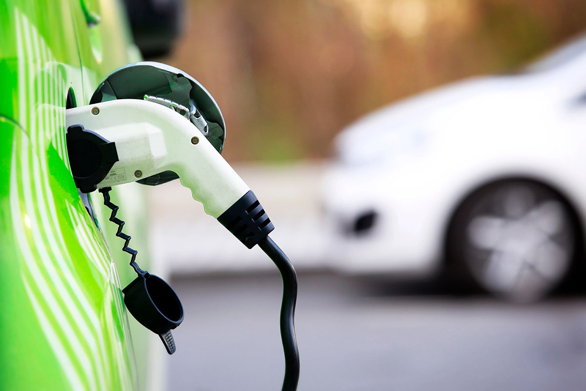 A close-up of a green electric vehicle with a charging cable plugged into its charging port, in the background a blurred white car is visible.