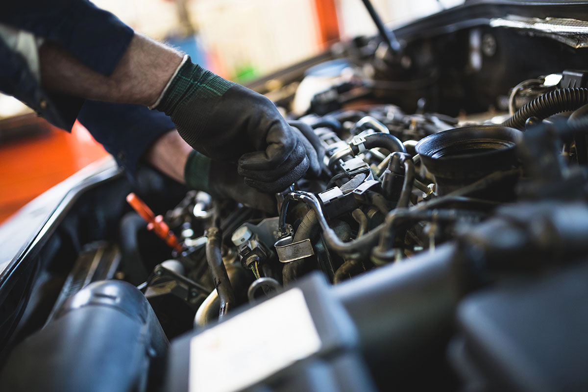 A mechanic wearing gloves is working on an automobile engine, using a wrench to tighten or adjust various components among the complex network of wires and tubes.