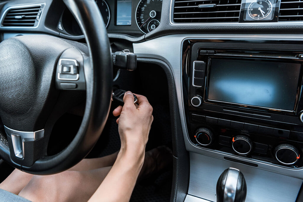 A close-up view of a person's hand inserting a car key into the ignition of a vehicle, with the steering wheel and dashboard controls visible in the background.
