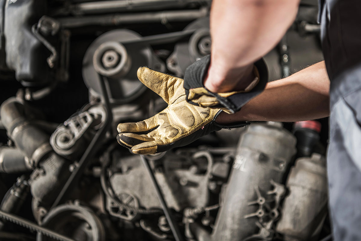 A mechanic wearing a yellow work glove adjusts their grip as they work on a vehicle engine, with various mechanical components visible in the background.