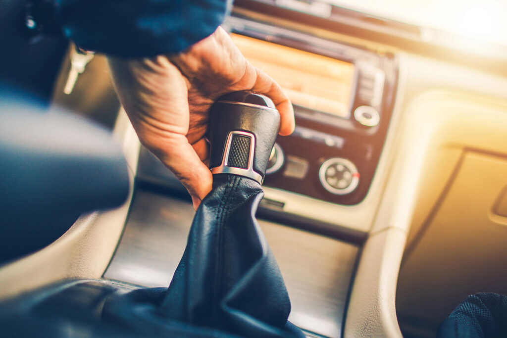 A close-up of a hand gripping a car's gear lever, with a blurred dashboard and infotainment system in the background.