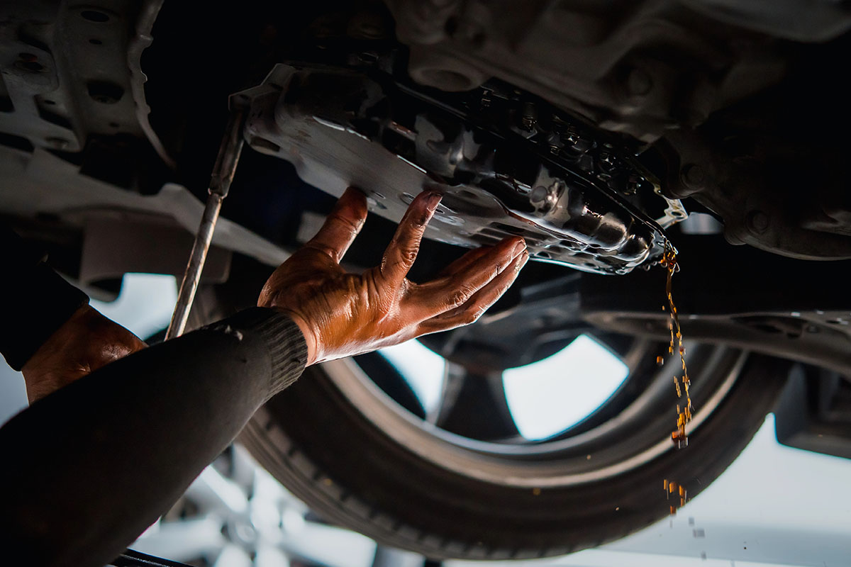 A mechanic's hand reaching up to remove a car oil pan under a vehicle, with oil dripping from the pan.