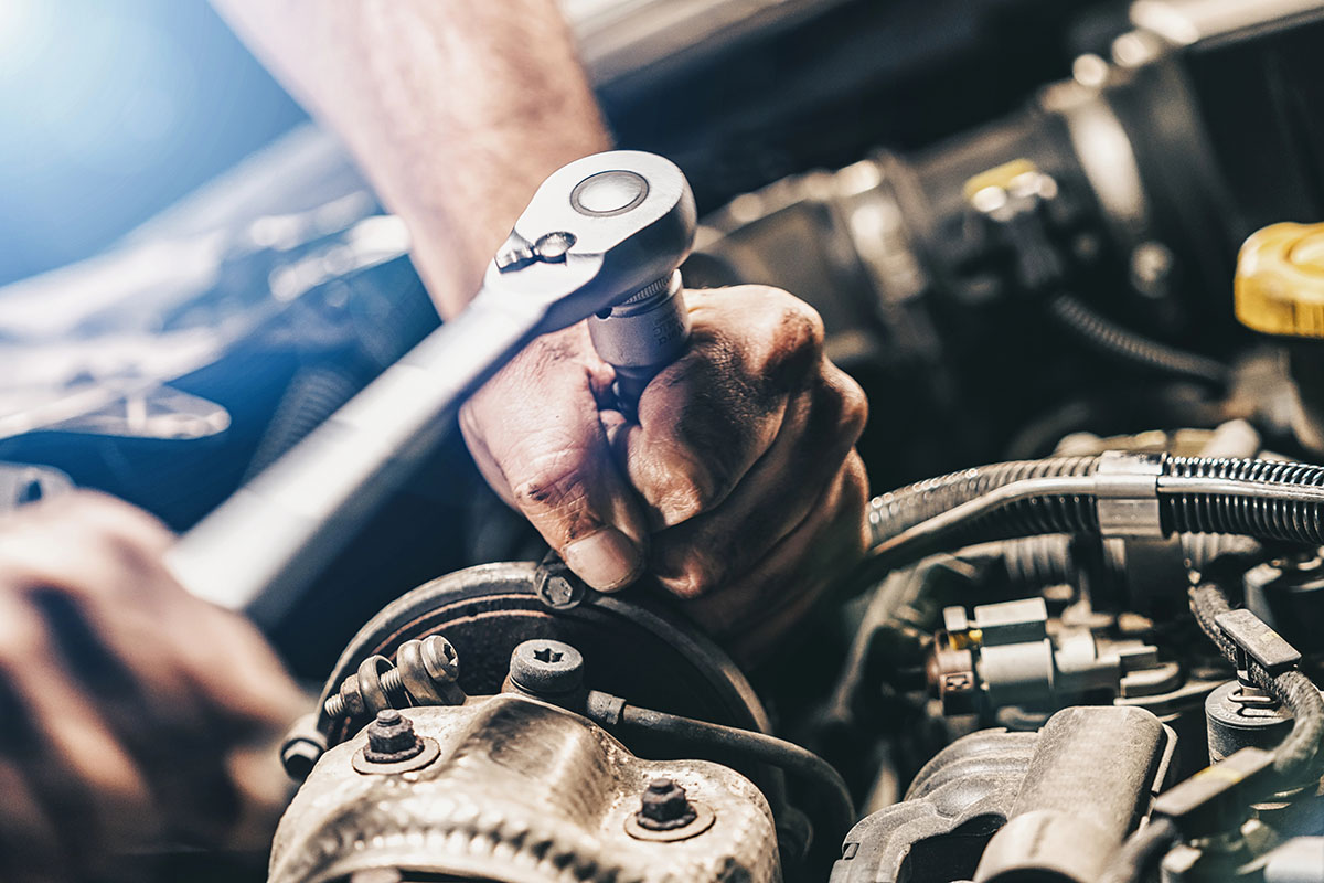 A close-up of a mechanic's hand using a ratchet wrench to work on an engine, surrounded by various automotive components and tools.