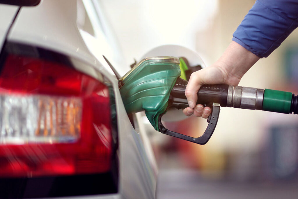 A hand holding a green fuel nozzle is shown inserting it into the gas tank of a car, with the car's rear and taillight partially visible in the background.