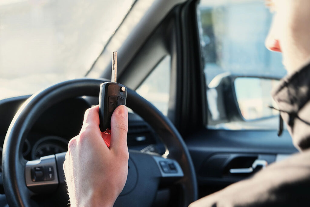 A person holding a car key fob in front of a steering wheel inside a vehicle, with sunlight streaming in through the window.