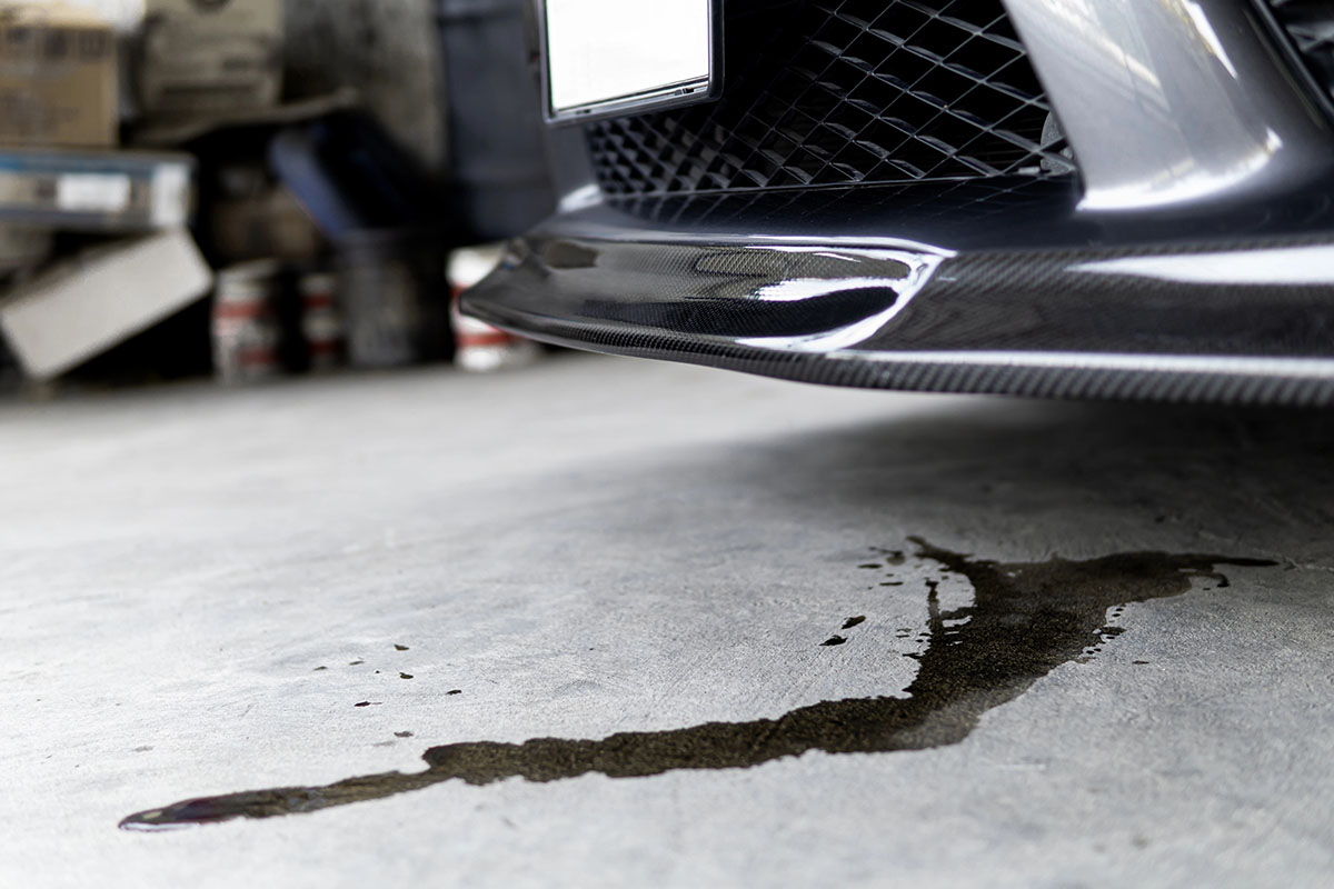 A close-up of a car's front bumper featuring a carbon fiber lip, with a dark stain of engine oil or fluid on a concrete floor underneath.