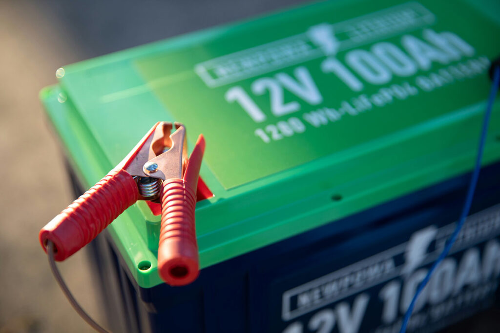Close-up of a green and blue battery with a label indicating "12V 100Ah" and a red jumper cable clamp resting on top.
