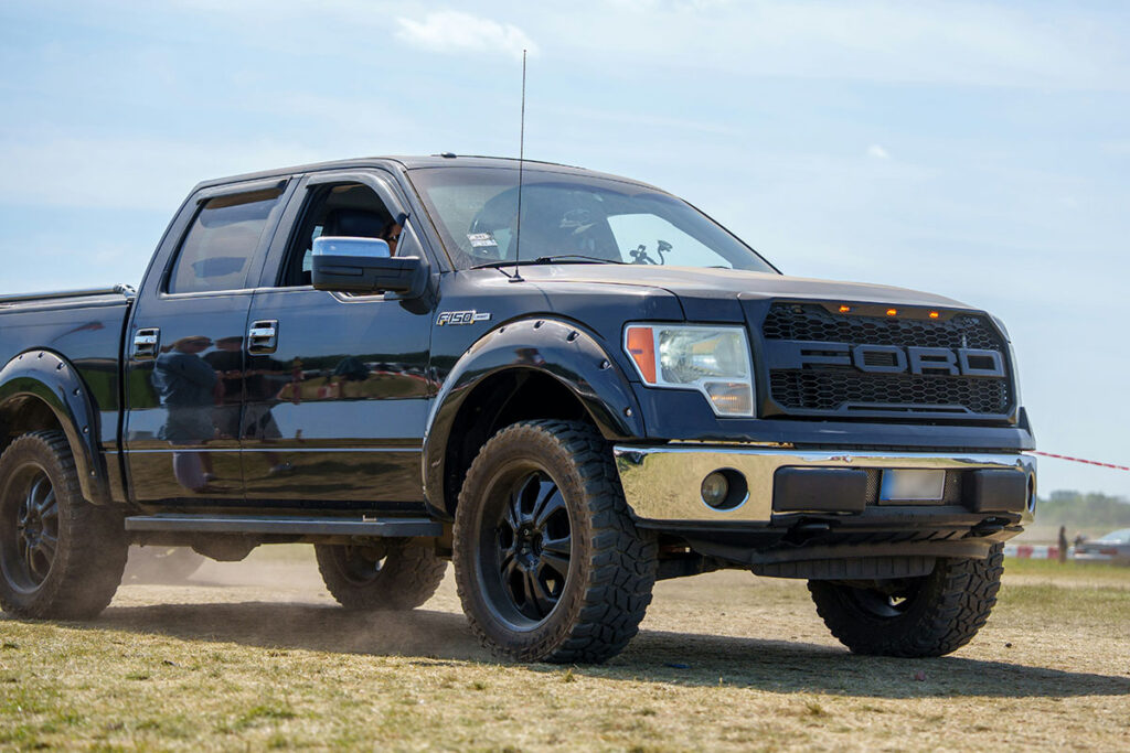 A black Ford F-150 pickup truck drives on a dirt field, kicking up dust, with a person visible in the driver's seat and a clear blue sky in the background.