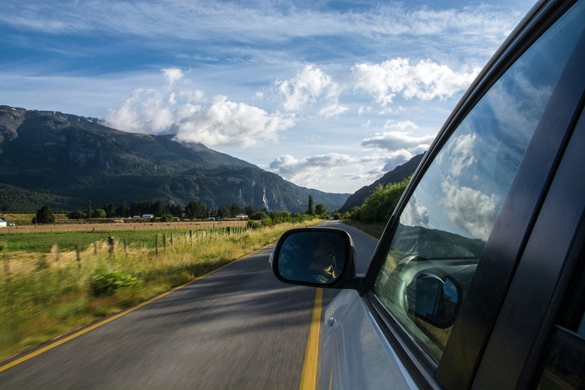 A view from the passenger side of a car, with mountains in the background and a scenic road lined with greenery and fields. The sky is partially cloudy, enhancing the picturesque landscape.