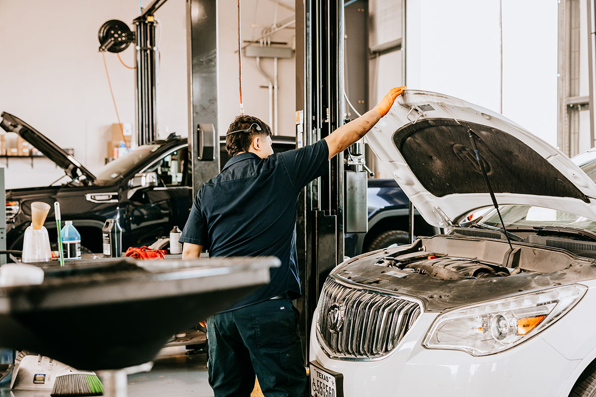A mechanic in a dark uniform inspects the engine compartment of a white vehicle with the hood raised in an auto repair shop, surrounded by tools and another car in the background.