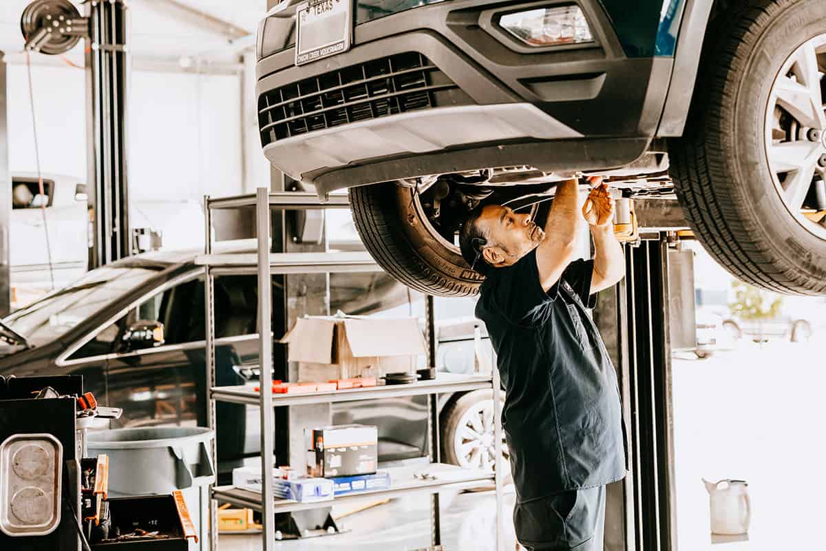 A mechanic works under a raised vehicle in an automotive shop, using a tool to make adjustments, with shelves of tools and supplies visible in the background.