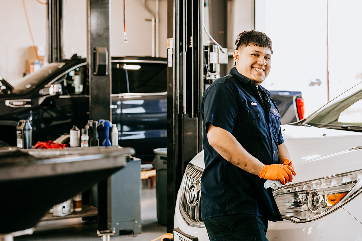 Photoshoot A technician in an automotive workshop smiles while holding a cloth and standing next to a white vehicle, with tools and cars in the background. He is wearing orange gloves and a dark uniform.