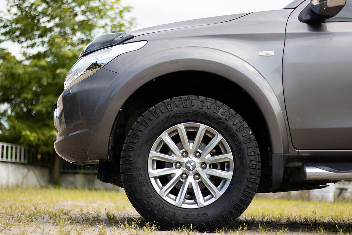A close-up view of the front tire and wheel of a gray sports utility vehicle (SUV) parked on grassy ground, with blurred greenery in the background.