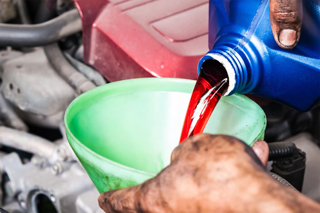A person pours red fluid from a blue container into a green funnel positioned over an engine. The hand shows signs of grease and dirt, indicating mechanical work.