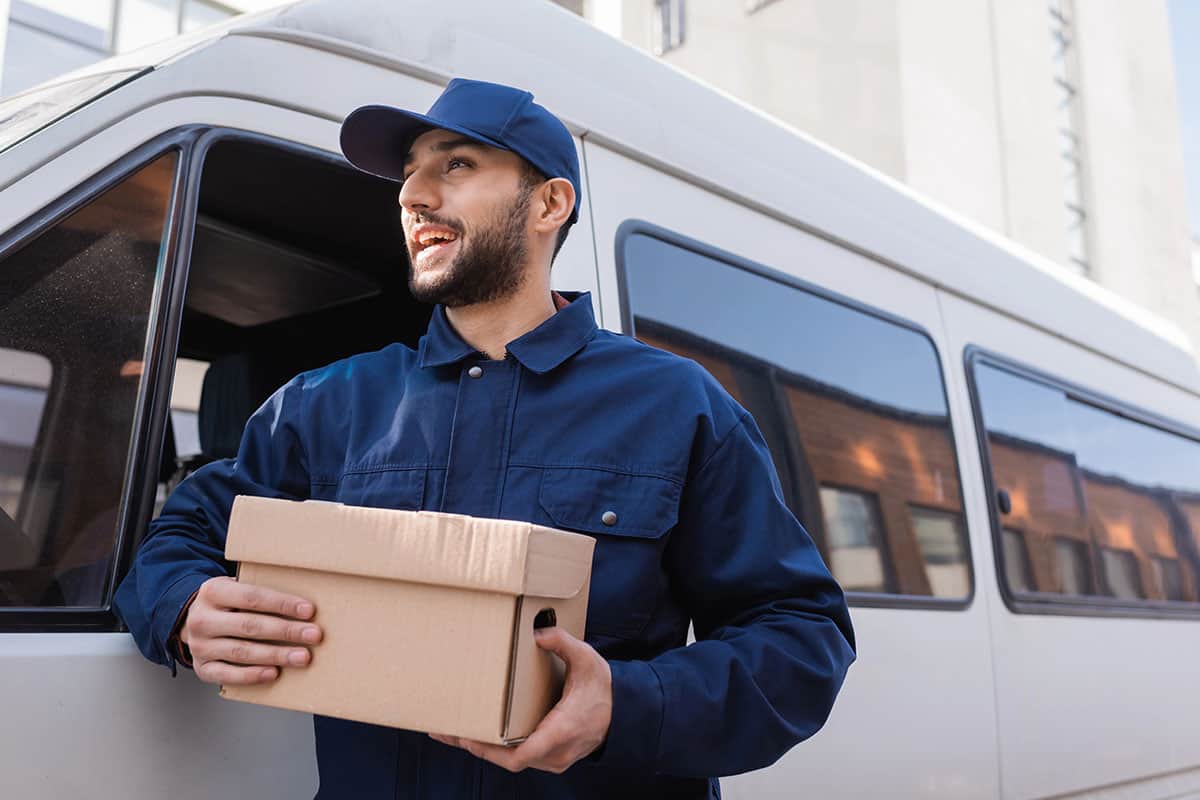 Delivery Driver A smiling delivery driver is holding a cardboard box while standing next to a commercial vehicle.