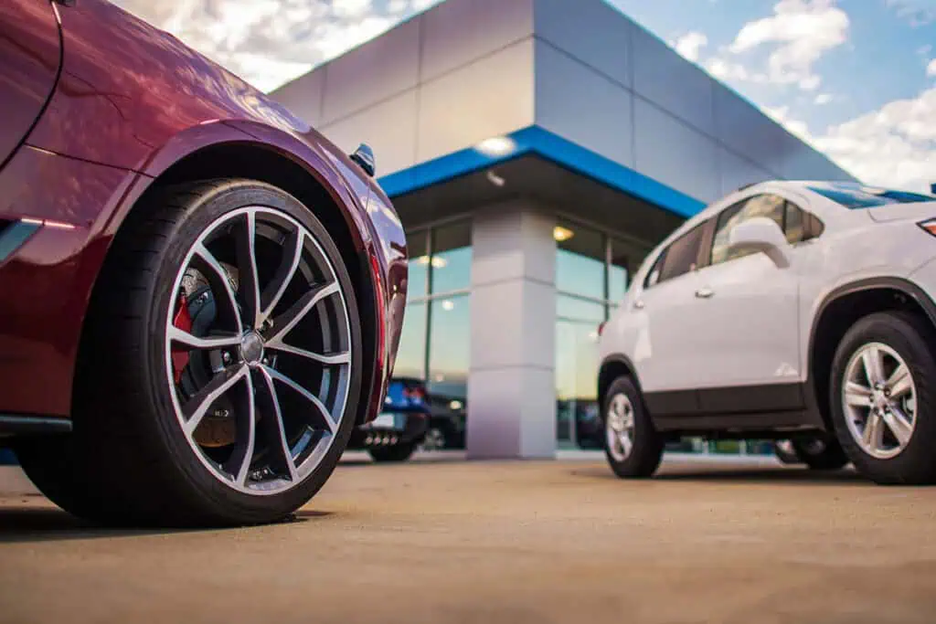 A low-angle shot of a maroon car's wheel in the foreground with a white SUV parked outside a modern car dealership showroom under a bright sky.